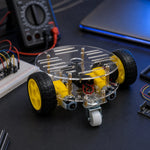 2WD robot car chassis with yellow wheels and clear acrylic base on a workbench, caster wheel visible underneath.