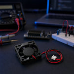 A 40mm 5V mini DC cooling fan with its red-and-black 2-pin JST wire lead, resting on a dark electronics workbench alongside a breadboard, multimeter, and jumper wires