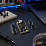 ESP32-C3 SuperMini board with its loose header pins in sharp focus on a dark electronics workbench with a breadboard, jumper wires, a multimeter, and a laptop softly out of focus.