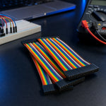 Rainbow ribbon jumper wires on a dark electronics workbench beside a breadboard and multimeter.
