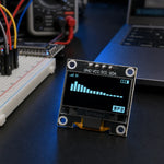 0.96" I2C OLED display module in sharp focus on a dark electronics workbench with a breadboard, jumper wires, a multimeter, and a laptop softly out of focus in the background.