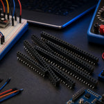 Breakable 2.54mm pin header strips on an electronics workbench with jumper wires, a breadboard, and a multimeter.