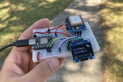Breadboard held outdoors with ESP32-C3 SuperMini, NEO-6M GPS module, and OLED display showing GPS coordinates