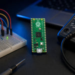 A Raspberry Pi Pico RP2040 microcontroller board standing on a dark workbench alongside a breadboard, jumper wires, and a multimeter