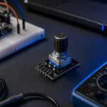 Rotary encoder module with knob fitted, on a workbench beside a breadboard and multimeter, illustrating its use as a physical menu/volume control.