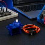 SG90 micro servo motor with its orange 3-pin connector on a dark workbench, with a breadboard, microcontroller, laptop, and multimeter in soft-focus behind it