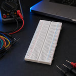 White 830-point solderless breadboard on a dark electronics workbench with jumper wires and a multimeter.