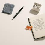 Real TCS3200 color recognition sensor composited onto a minimal bright white desk scene with anti-static bags, a mechanical pencil, a notebook with a hand-drawn PCB sketch, and wireless earbuds in their case.