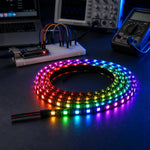 Coiled WS2812B addressable LED strip glowing in rainbow colors on a workbench with a breadboard, dev board, oscilloscope, and multimeter in the background.